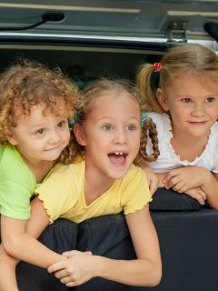 three happy kids with blonde hair in car looking out of trunk from back seat