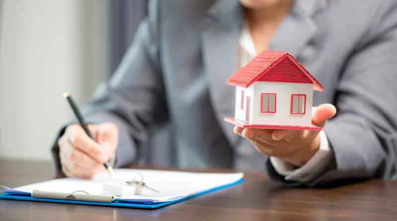 woman holding model house in one hand and signing a document with the other