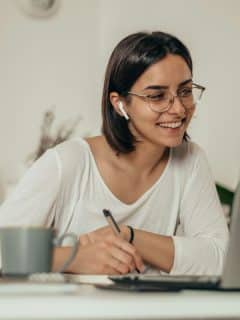 woman in white top working at home with laptop and writing in notebook