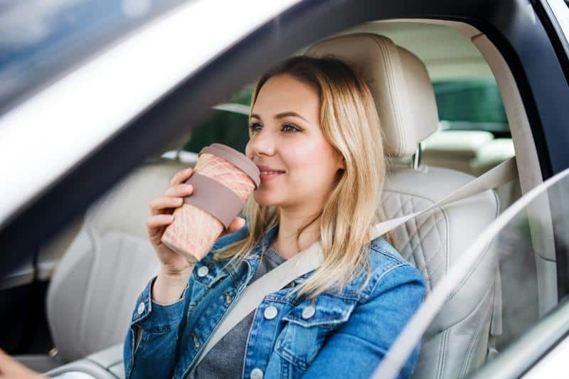 blonde woman in denim jacket buckled into car holding cup of coffee