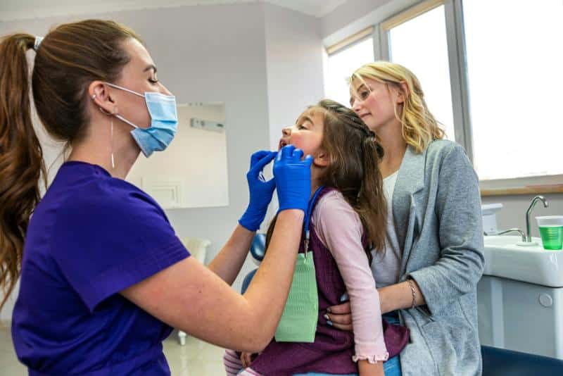 female dentist looking at little girl's teeth while mom supports