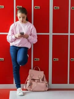 preteen girl with cell phone standing against red lockers wearing a pink shirt and jeans with pink backpack on floor beside her