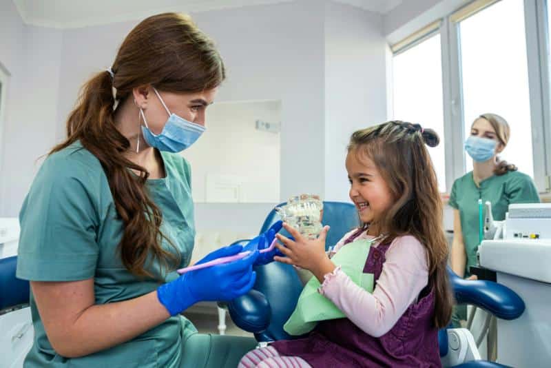 little girl smiling while looking at model of teeth dentist is showing her