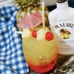 Gorgeous layered drink called a Malibu Sunset in glass with pineapple and cherry garnish and small umbrella