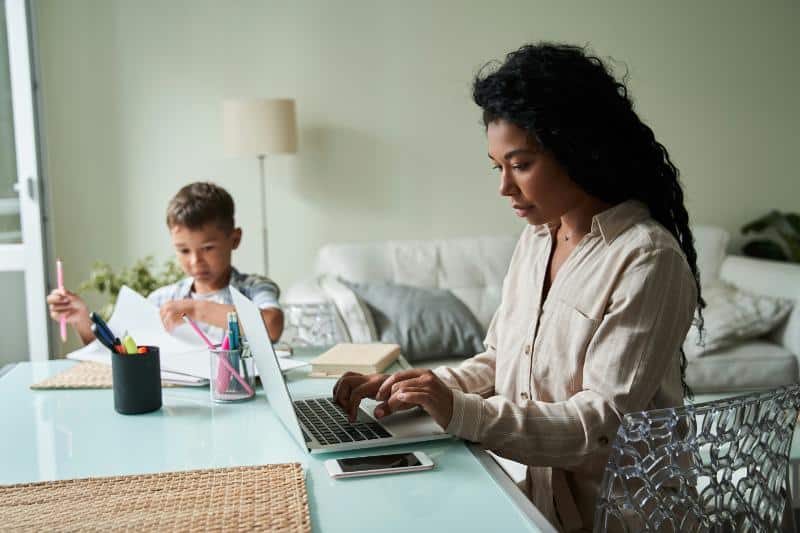 mother working on laptop while son colors at table beside her