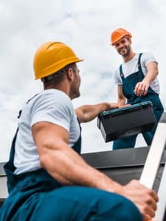 two men working on a roof, one reaching a toolbox to the other