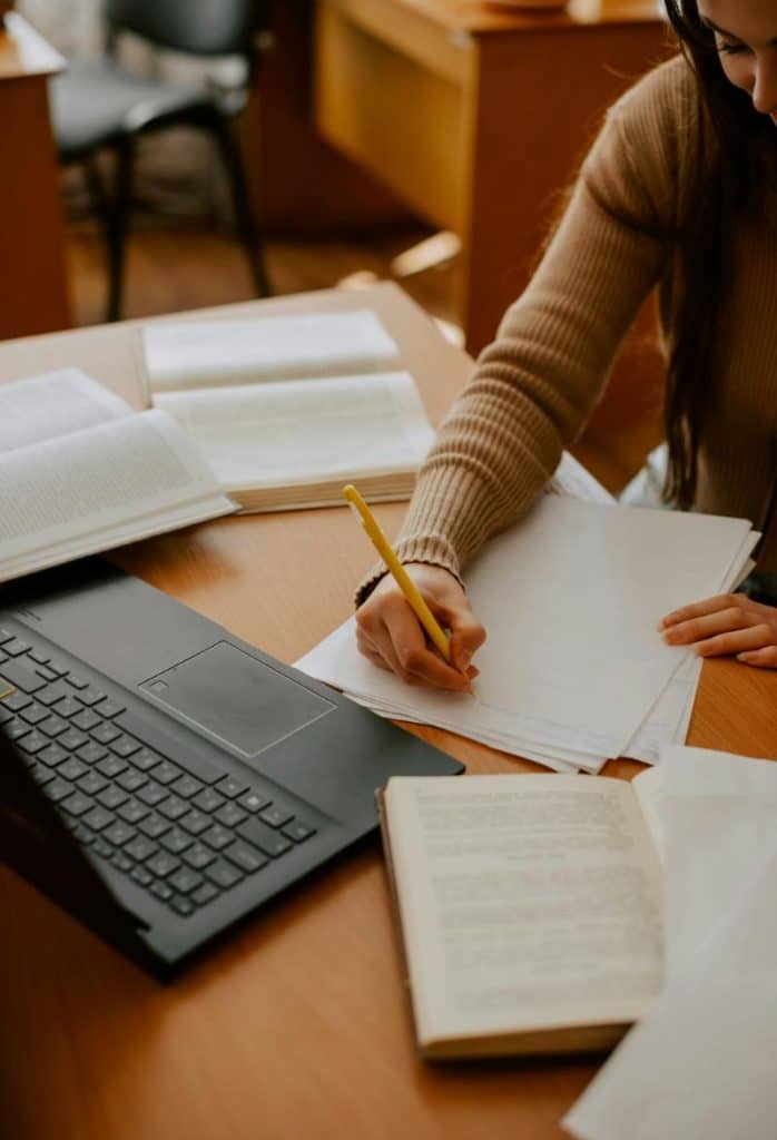 woman writing on paper surrounded by books and a laptop