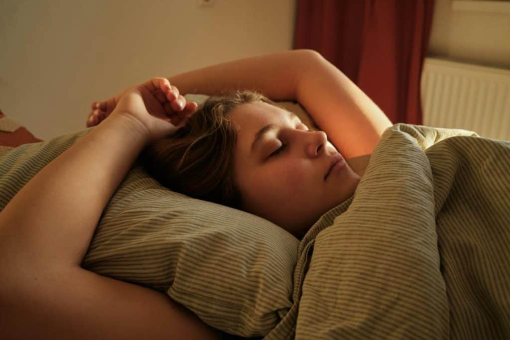 woman in bed with eyes closed, sunlight starts to stream in through windows