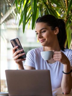 woman holding a cup of coffee and looking at her phone, laptop on table in front of her