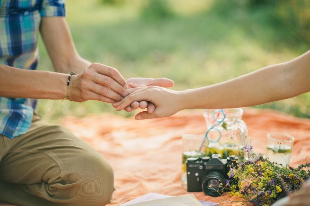 man and woman on picnic blanket, man putting ring on woman's hand (neither face is shown)