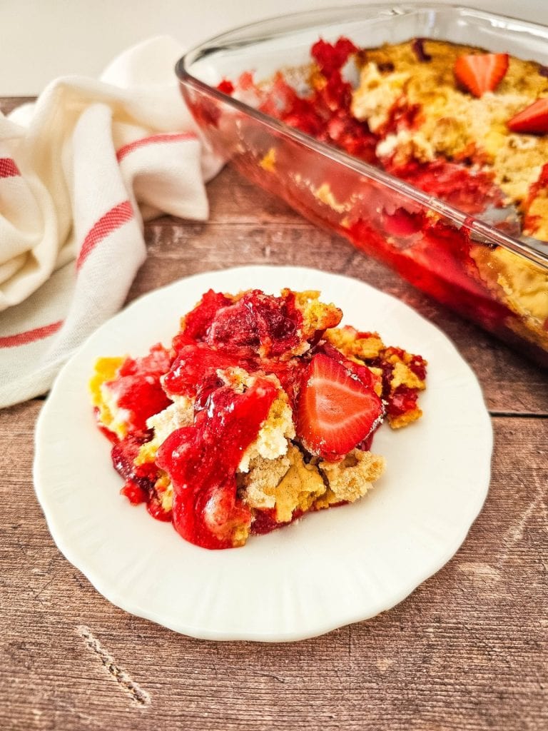 a piece of strawberry cheeesecake dump cake on a white plate with the rest in a pan behind