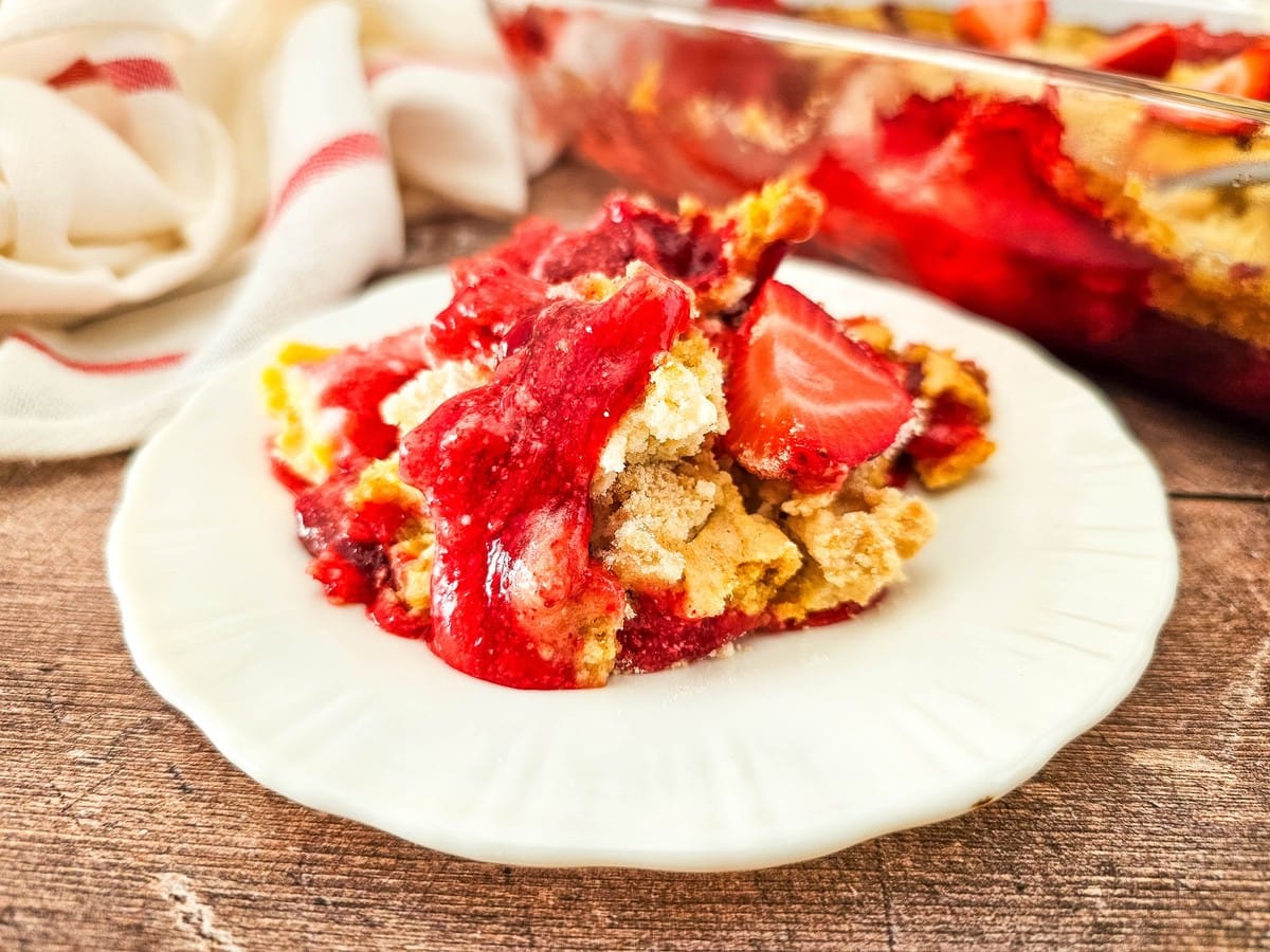close-up of strawberry cheesecake dump cake on white plate with baking dish behind