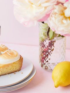 slice of lemon meringue cake on white dish with a vase of flowers and fresh lemons off to the side