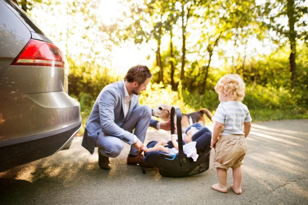 dad with toddler, baby, and dog beside car