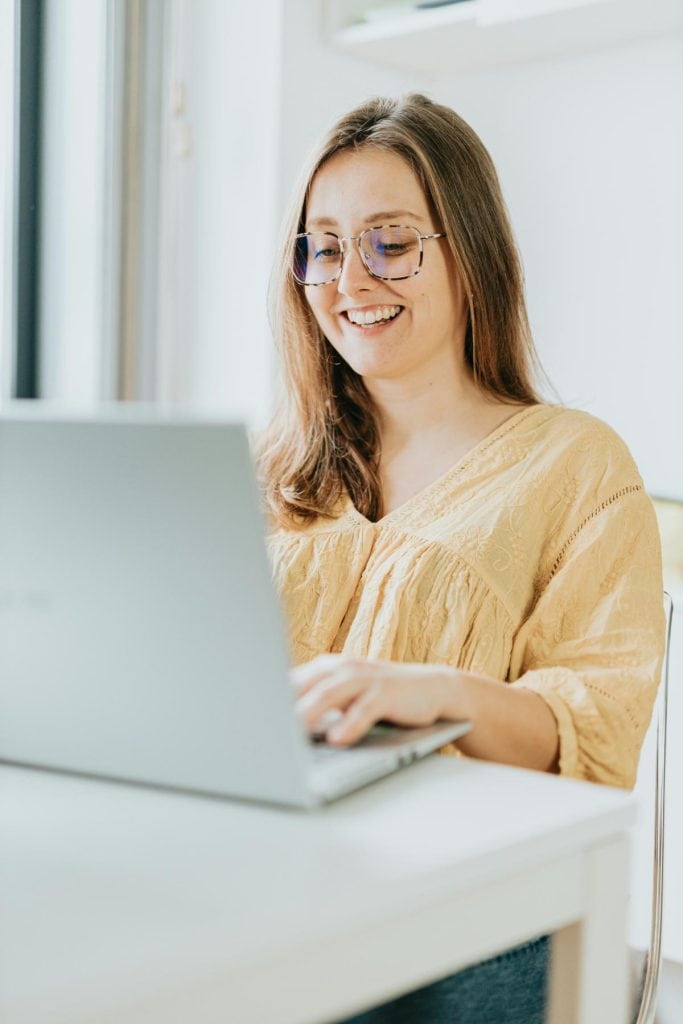 woman with glasses smiling while sitting at a desk and using a laptop