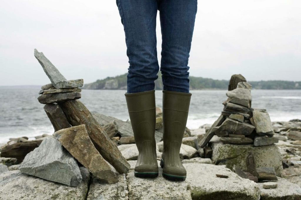 person in rain boots standing on rocks near water