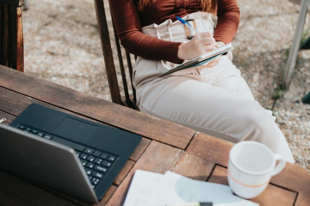 woman sitting at a table with a laptop writing in a notebook