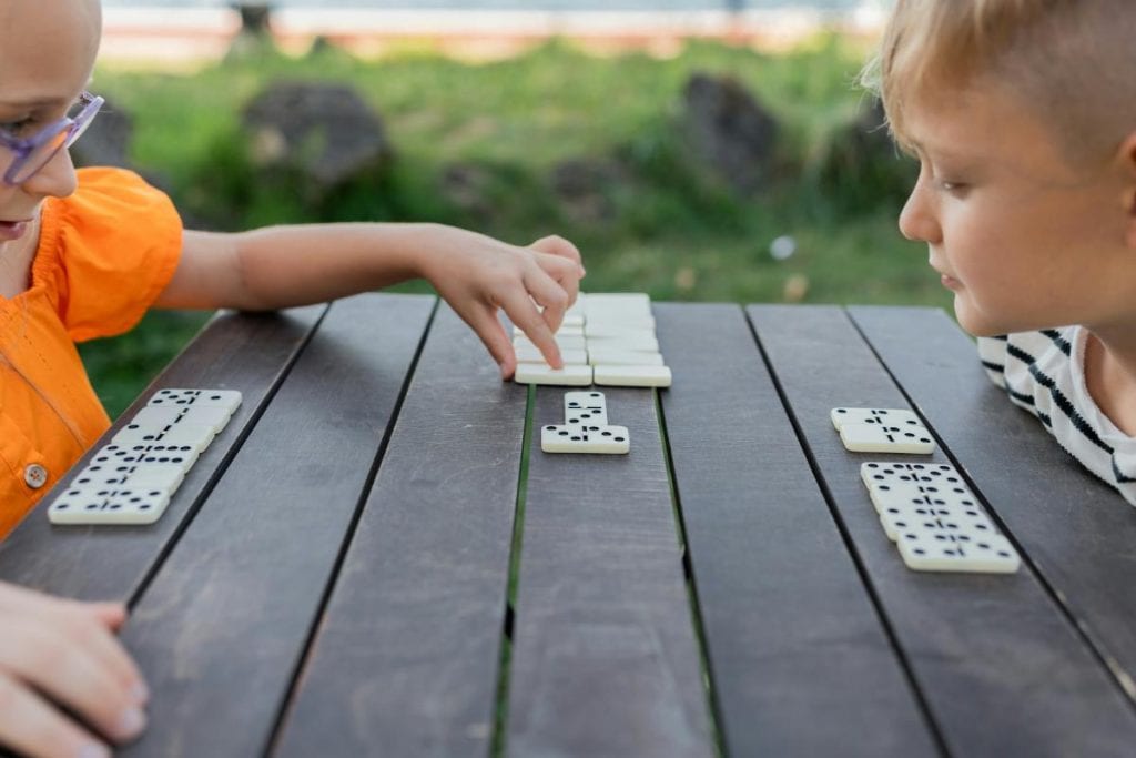 kids playing with dominos outdoors at a wooden table