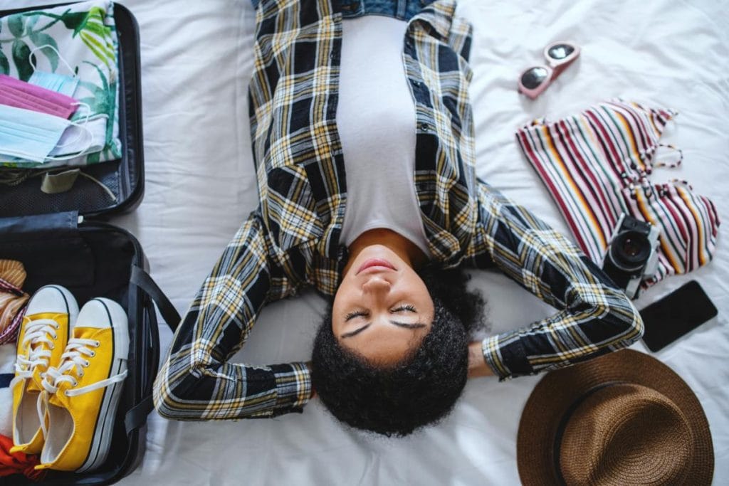 young woman laying in bed with suitcase and travel items surrounding her