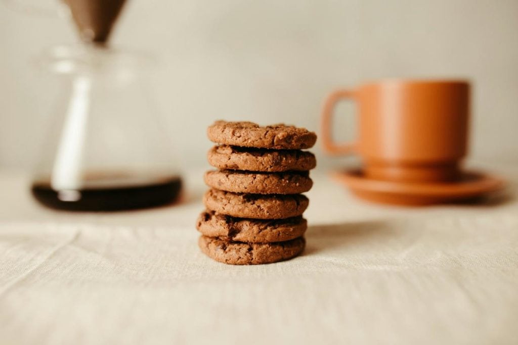 a stack of chocolate cookies with a cup of coffee and coffee pot in background