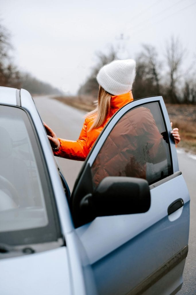 woman in orange coat standing beside car with door open looking behind her