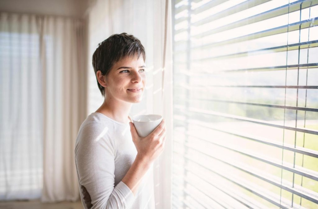 woman looking through blinds enjoying a cup of coffee