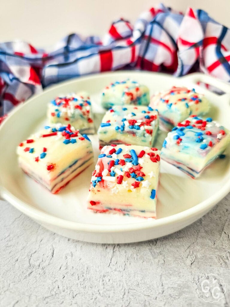 Close-up of creamy red, white, and blue fudge squares topped with star sprinkles, served on a white dish with a patriotic plaid cloth in the background—perfect for Fourth of July or Memorial Day desserts.