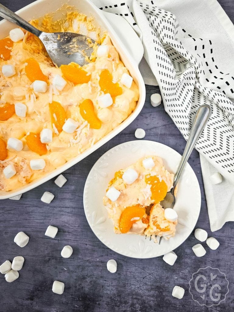 Serving of mandarin orange jello salad on a white plate with a fork, surrounded by scattered mini marshmallows.