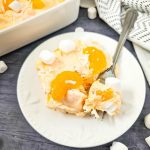 A spoonful of fluffy orange jello salad with whipped topping, pudding, and mandarin oranges, scooped out of a casserole dish.