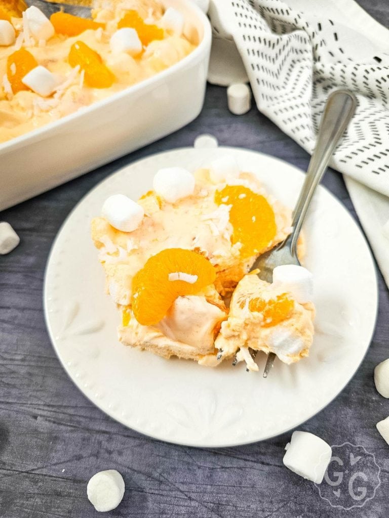 A spoonful of fluffy orange jello salad with whipped topping, pudding, and mandarin oranges, scooped out of a casserole dish.