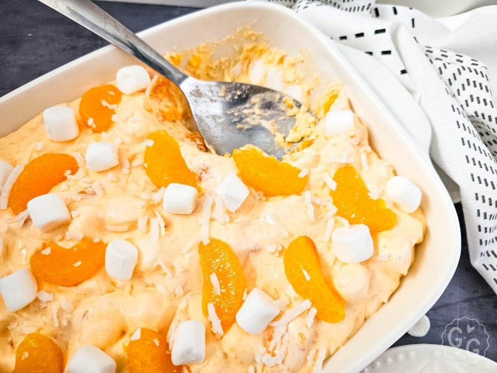 Mandarin orange fluff salad served in a white baking dish and on a white plate, styled with coconut and marshmallows for a retro no-bake dessert.