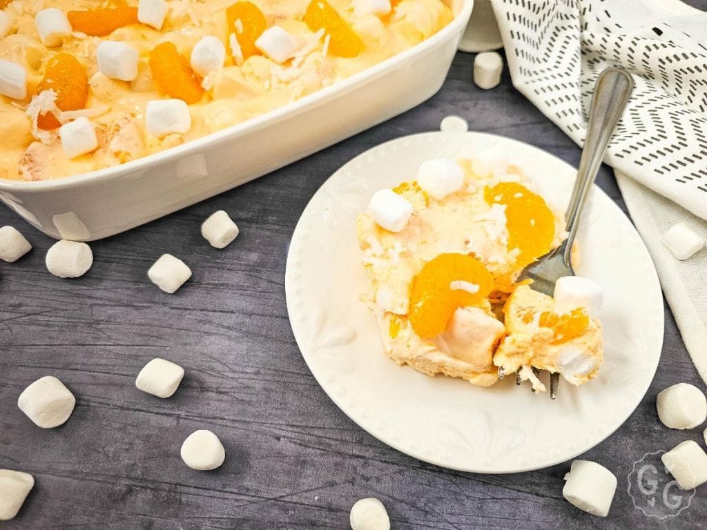 Vintage-style orange fluff salad with Cool Whip, pudding, and marshmallows in a white dish, partially served, with a patterned kitchen towel nearby.