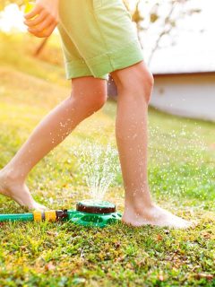 Child walking barefoot through backyard sprinklers—watering early in the morning helps maintain a healthy lawn during hot summer months.