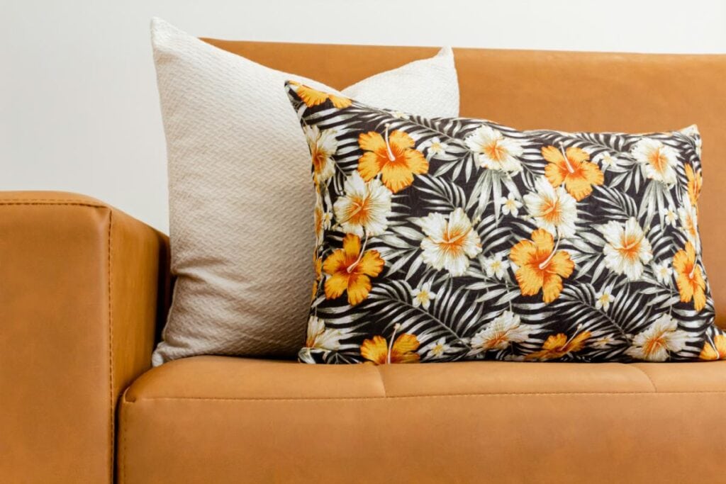 Tan leather sofa styled with a bold tropical floral pillow in black, orange, and white, paired with a textured cream accent pillow for a stylish living room look.