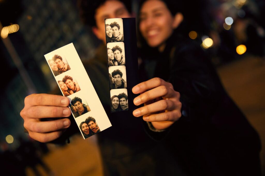 A smiling couple holding two strips of photo booth pictures, one in black and white and one in color, perfect as fun event keepsakes.