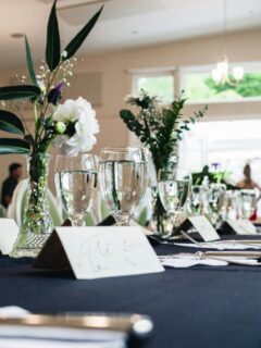 Elegant wedding table setup featuring clear wine glasses, floral centerpieces, black tablecloths, and handwritten place cards ready for guests.