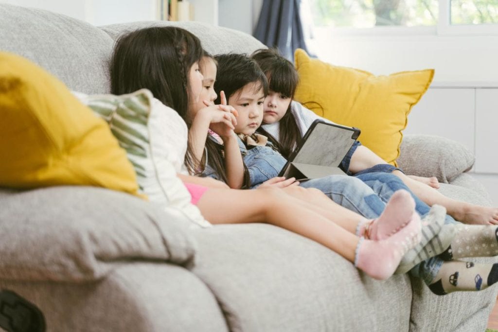 four kids sitting on the couch watching a tv show on a tablet device