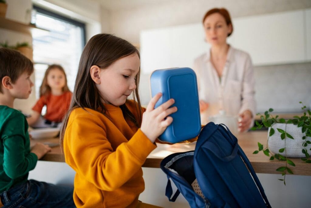 Elementary-aged girl in a yellow sweater packing a blue lunchbox into her backpack while getting ready for school at home.