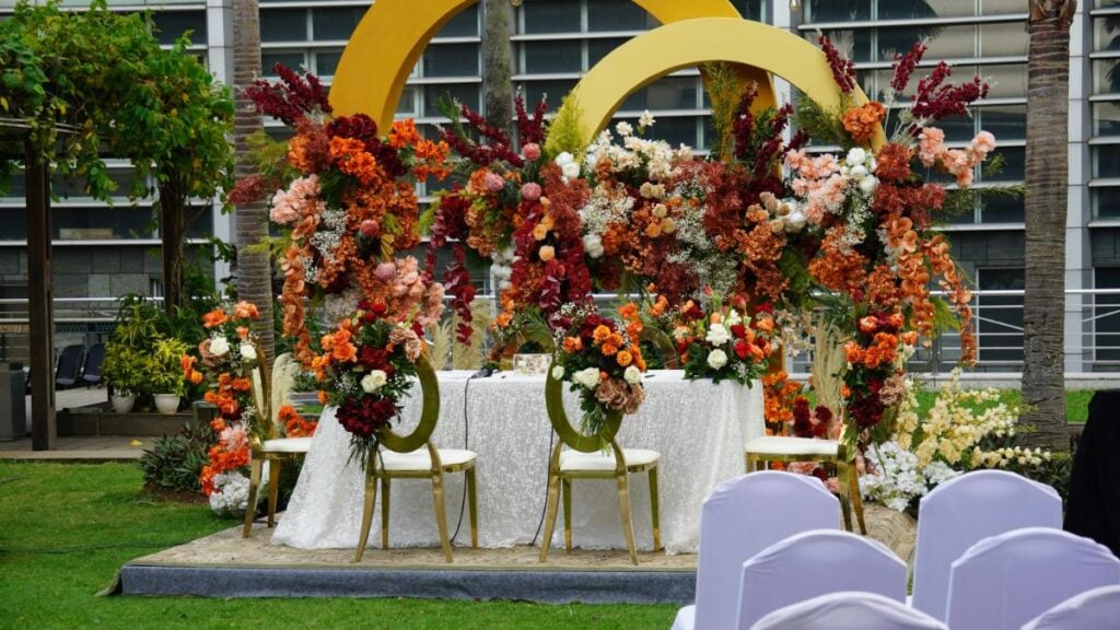 Outdoor head table decorated with vibrant floral arrangements in shades of red, orange, and cream, set against a modern building backdrop.