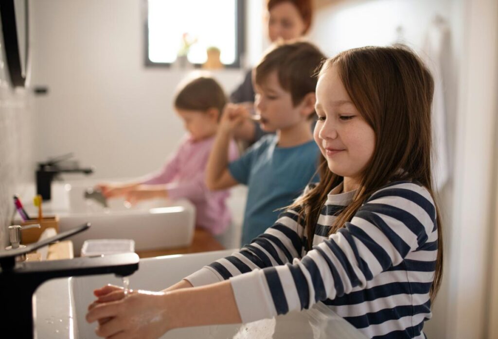 Young kids brushing teeth and washing hands at the sink as part of their morning school routine in a well-lit bathroom.