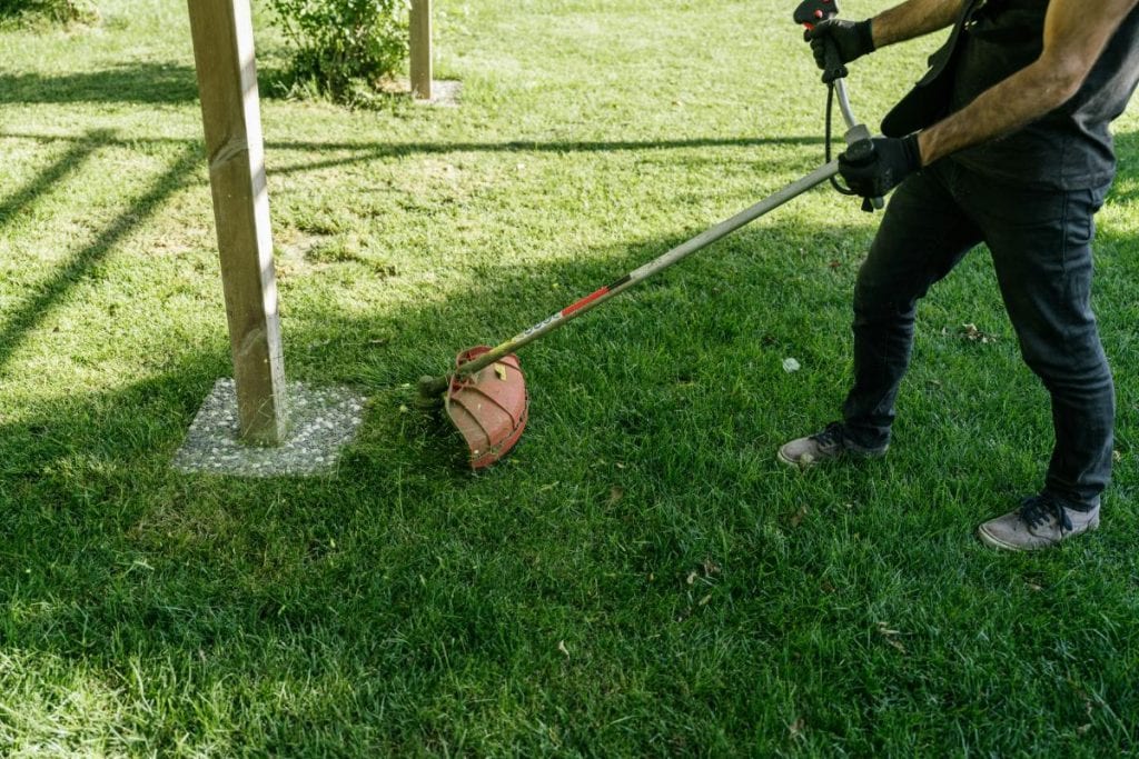 Person using a string trimmer to clean up grass along a post—trimming and edging add a polished look to your summer yard.