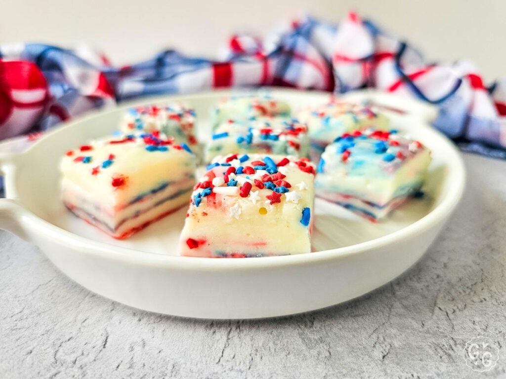 Side angle of rich and creamy red, white, and blue fudge stacked on a white serving platter, decorated with patriotic sprinkles for a classic summer treat.