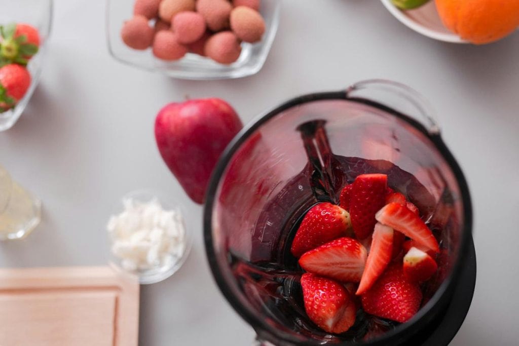 strawberries in a blender surrounded by other ingredients like an apple and nuts