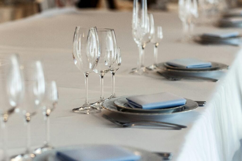 Close-up of a formal event table featuring silver charger plates, folded blue napkins, and multiple wine glasses on a crisp white tablecloth.