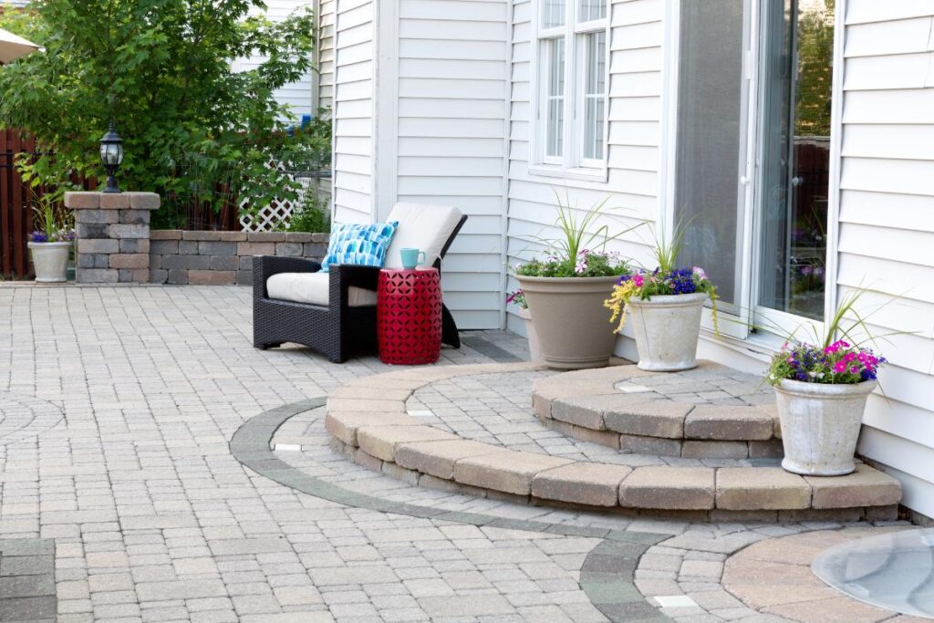 Bright outdoor patio space featuring a white vinyl-sided home, cozy black wicker chair with a blue pillow, red accent table, and multiple large planters filled with vibrant summer flowers and greenery.