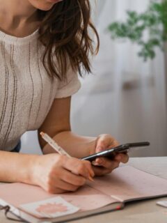 Woman sitting at a table planning her monthly schedule using a notebook and smartphone to practice time management and organization skills.