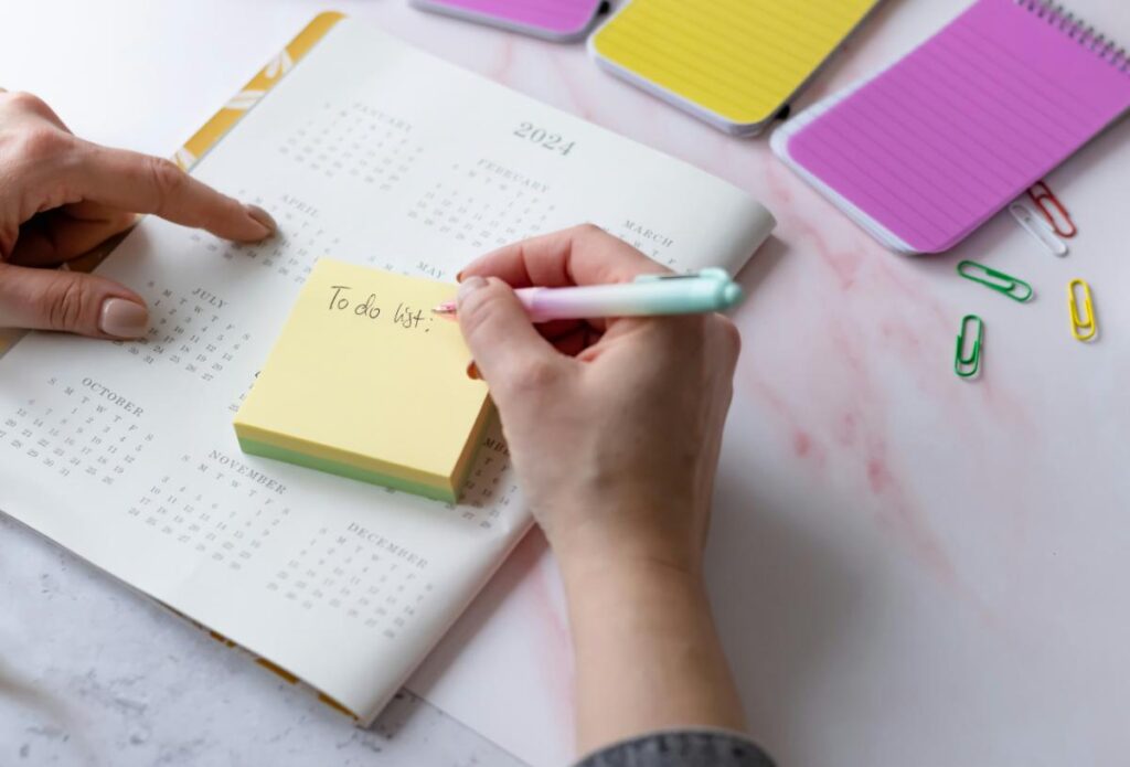 Hands writing a to-do list on a yellow sticky note placed on top of a yearly planner, showing effective time management and planning strategies.