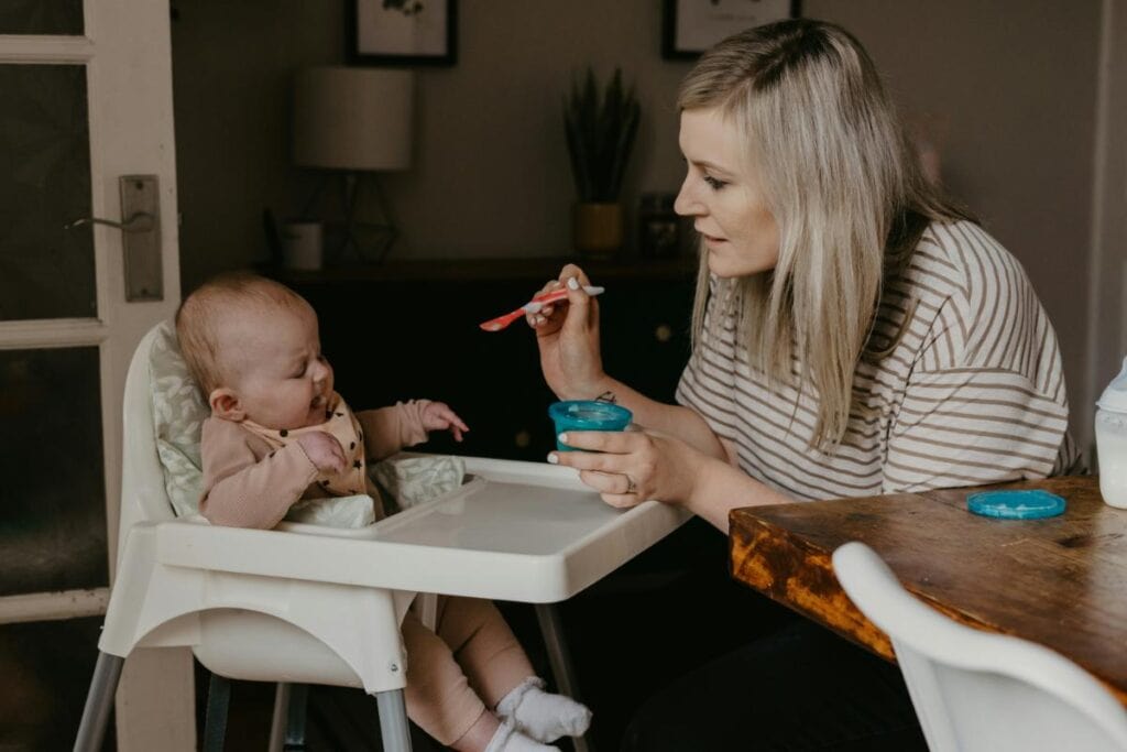 Mother feeding baby solid food in high chair with spoon; baby wearing bib and making a hesitant face during mealtime.
