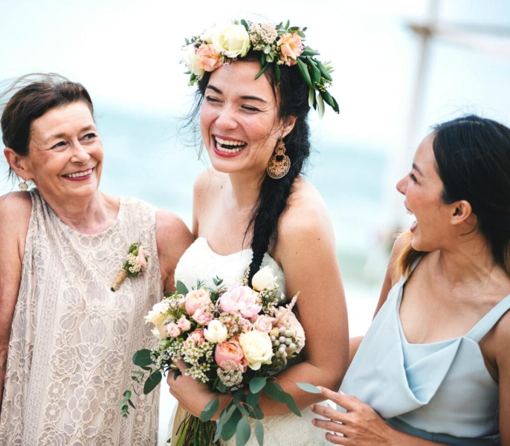 Bride laughing with her mother and bridesmaid on the beach, holding a pastel bouquet and wearing a floral crown, showing examples of flattering wedding party dresses.