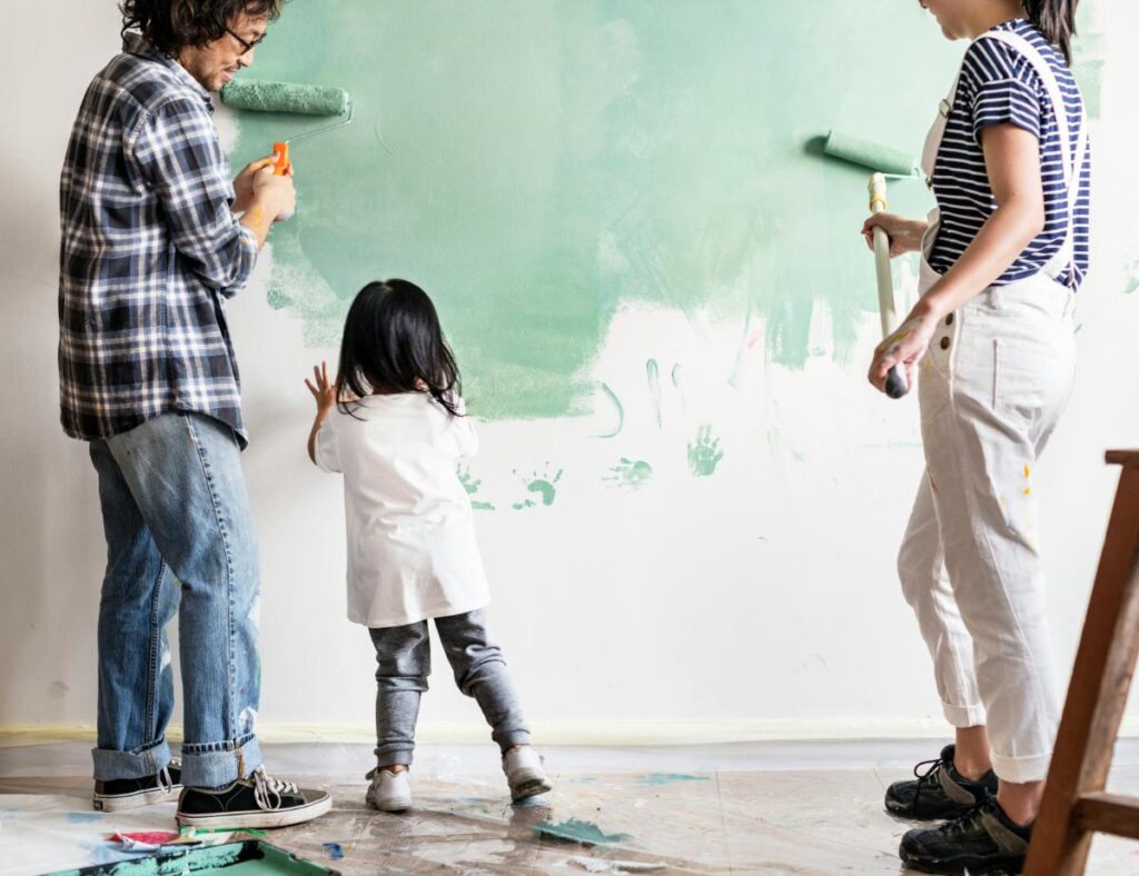 Family painting a green accent wall together with paint rollers and brushes during a DIY home decorating project.